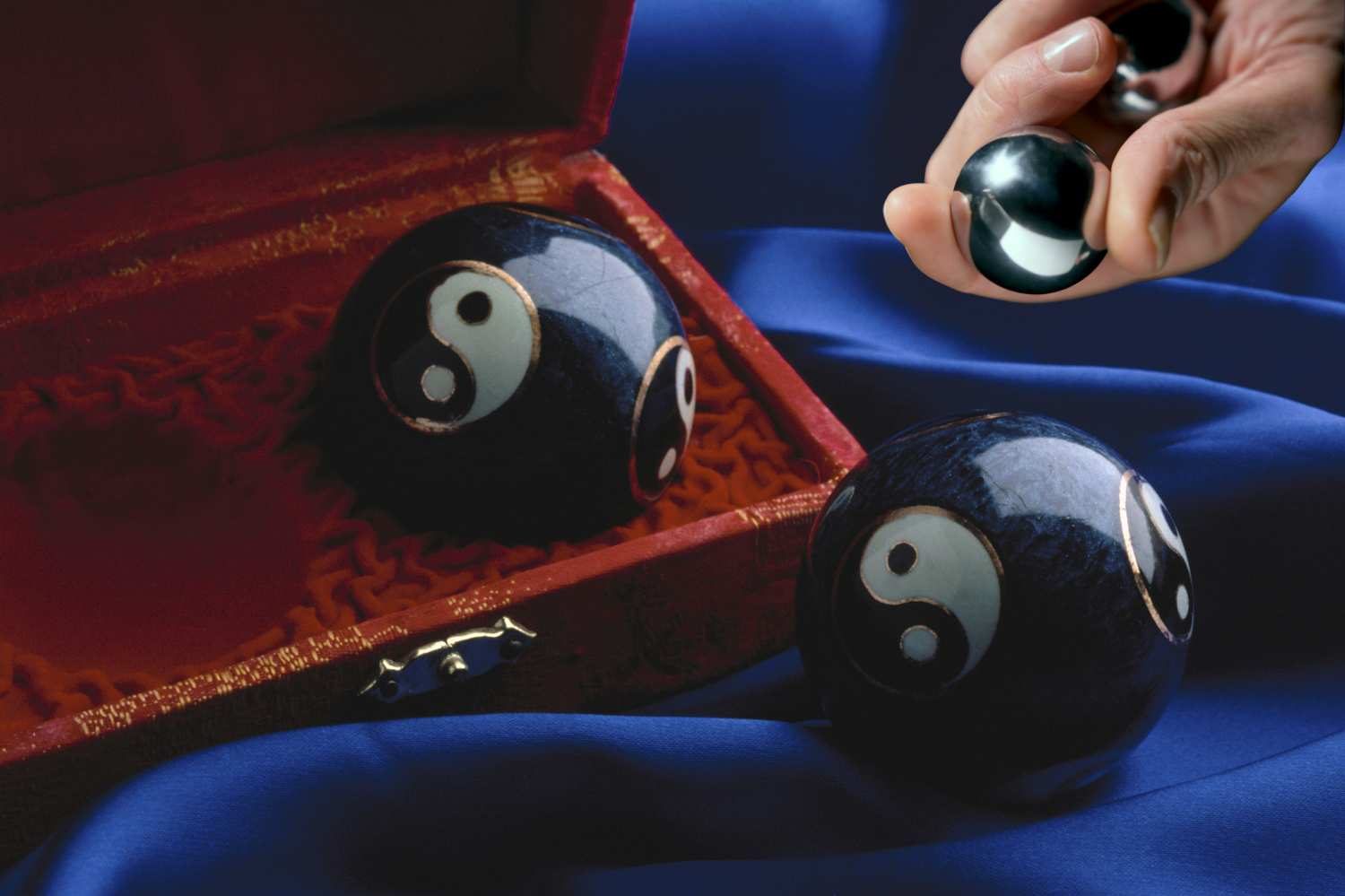 En hand håller i två hälsokulor i metall nära två yin-yang-hälsokulor på en blå sammetstyg.